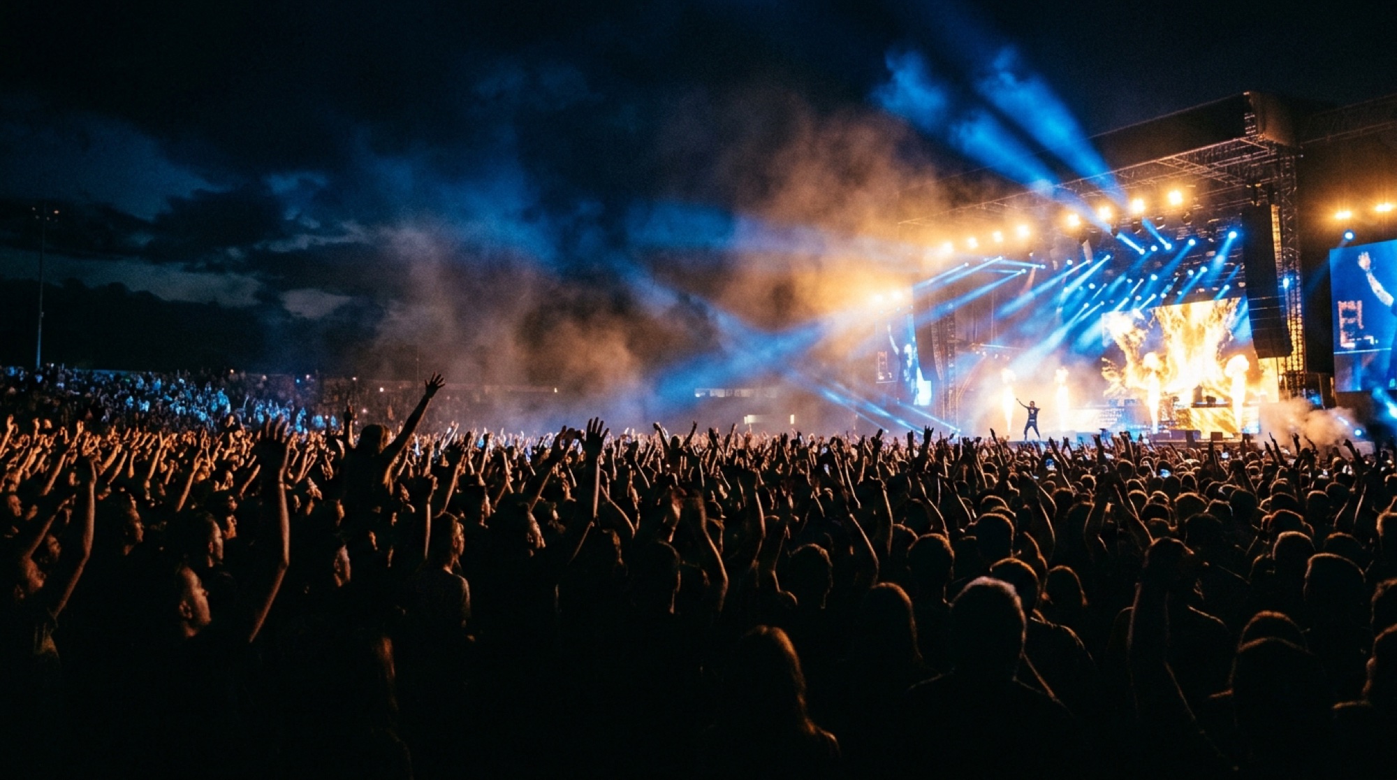 A festival crowd with hands in the air against the stage lights and haze